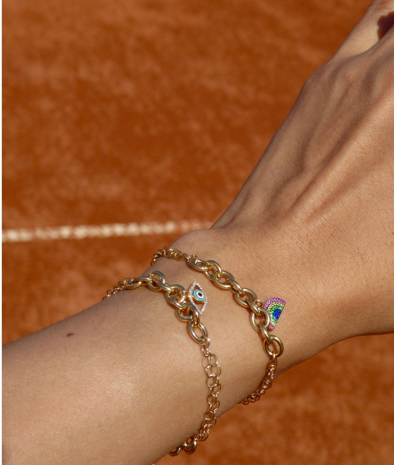 Gold chain bracelet with rainbow charm on wrist against a brown background.